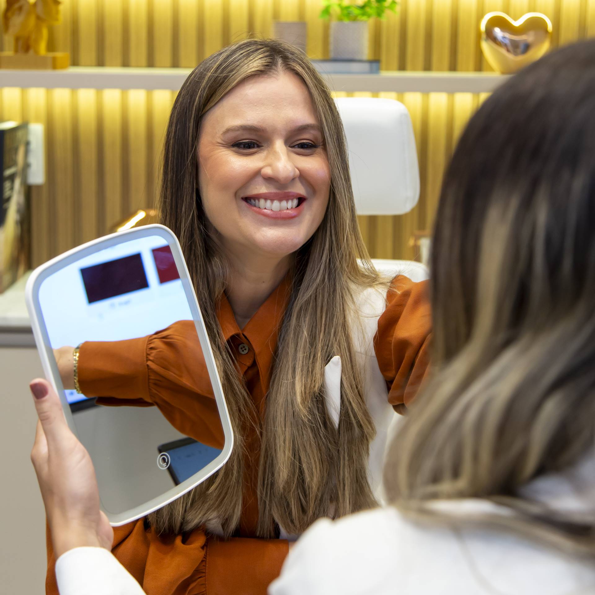 Dra. Thatiane Fernandes, enfermeira esteta da Revera Clinic, toca suavemente o rosto de uma paciente sorridente que segura um espelho, em um momento de cuidado, avaliação estética e valorização da autoestima.